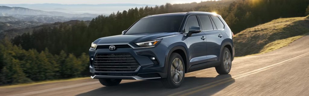 A blue Toyota Grand Highlander SUV driving on a scenic road surrounded by mountains and trees