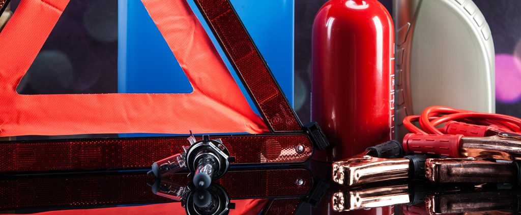 The image shows essential car accessories: a warning triangle, halogen bulb, fire extinguisher, and jumper cables.
